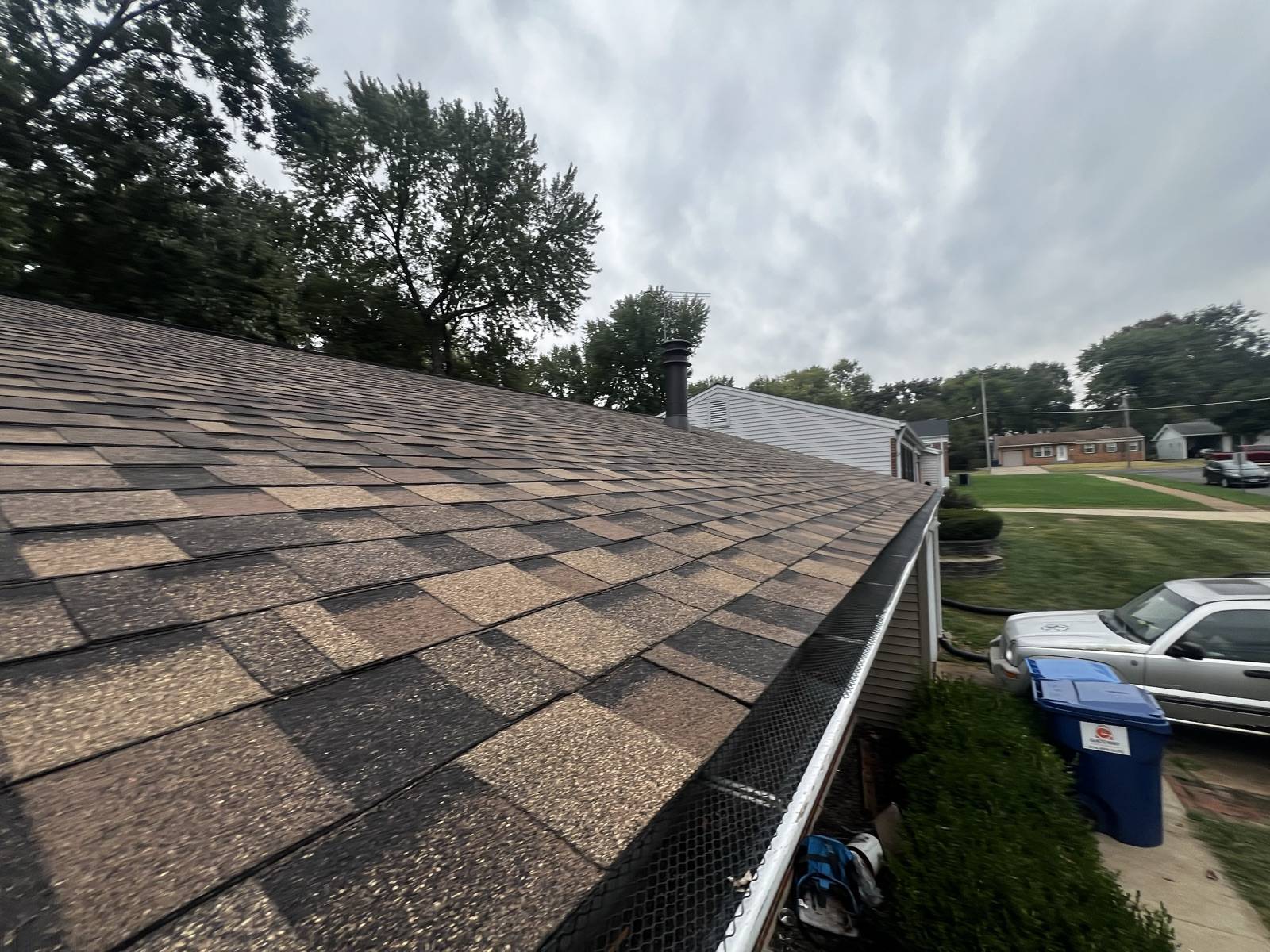 A brown shingle slope running down to a white gutter fitted with black mesh guard, photographed under a heavy overcast sky with full summer trees behind