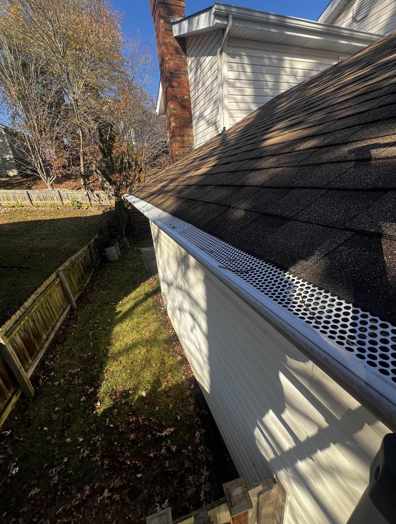 A white aluminium gutter fitted with a punched metal guard running along the eave of a dark shingle roof, with fallen leaves on the lawn below