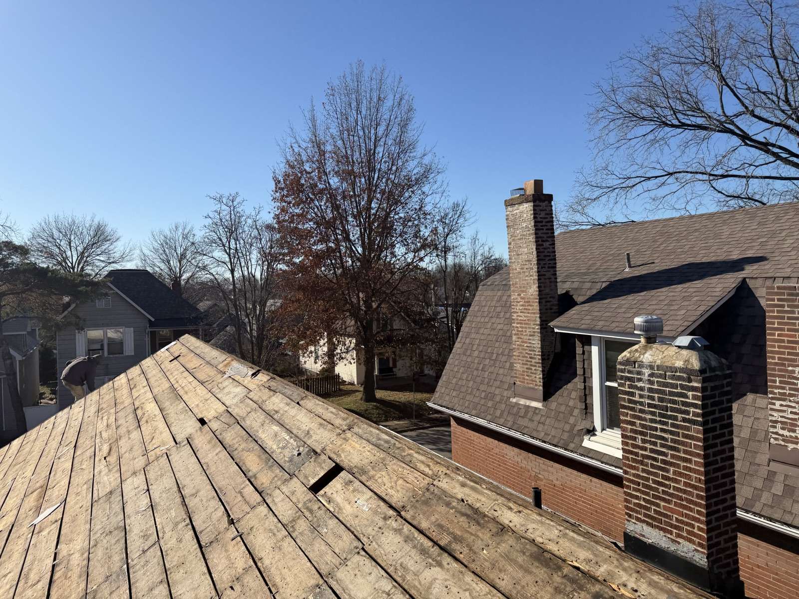 The hip of a roof stripped to bare boards, with the brick chimneys and shingled slopes of the same house beyond and bare winter trees over the neighbouring rooftops
