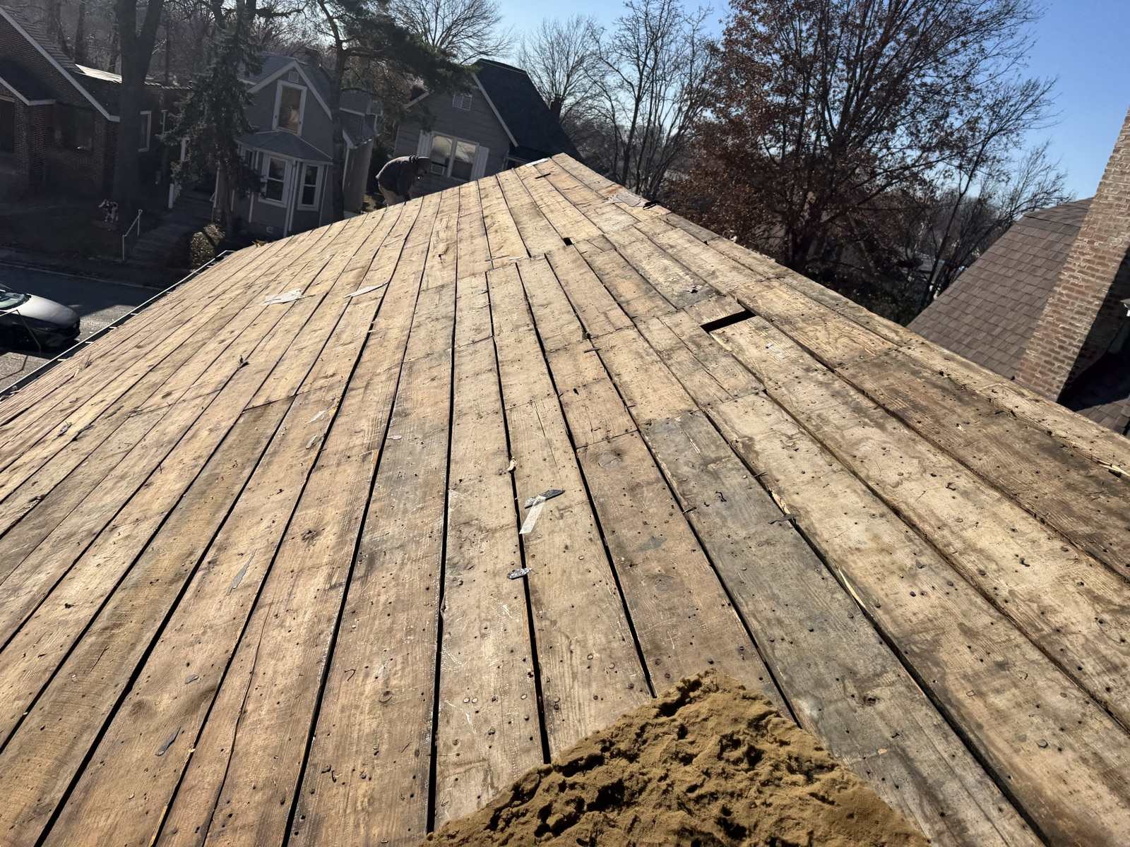 A steep roof slope stripped back to its original board decking, plank gaps and old nails showing, with a residential street and parked car below
