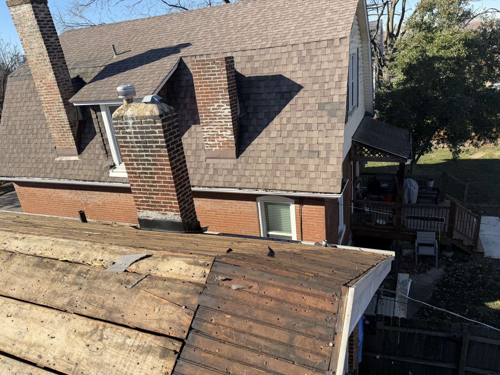 A lower porch roof stripped to dark board decking with scraps of old felt still attached, the main slope of the brick house above it still shingled