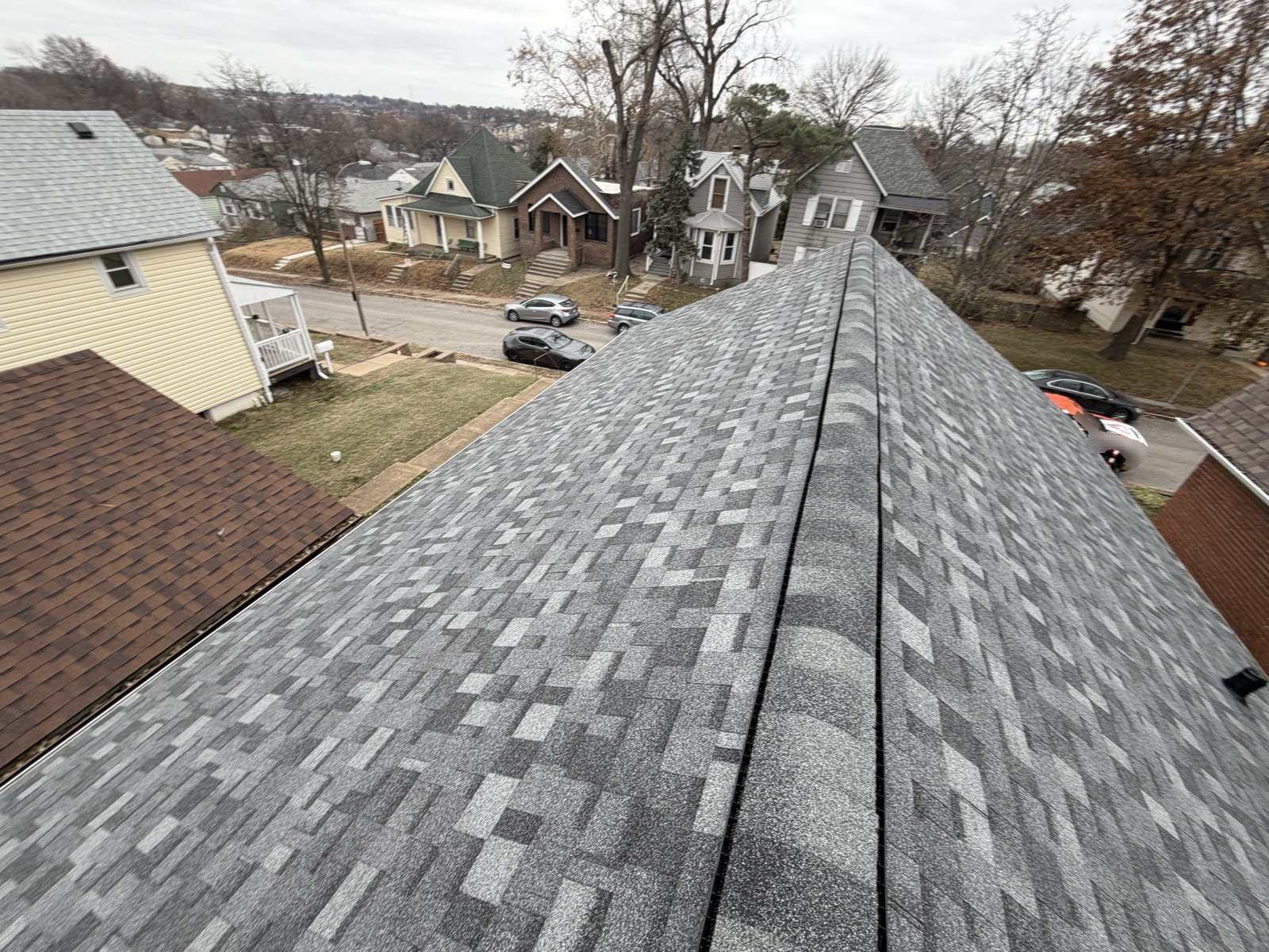A finished grey architectural shingle roof photographed along its ridge, looking down over an older St. Louis street of brick and frame houses under a flat winter sky