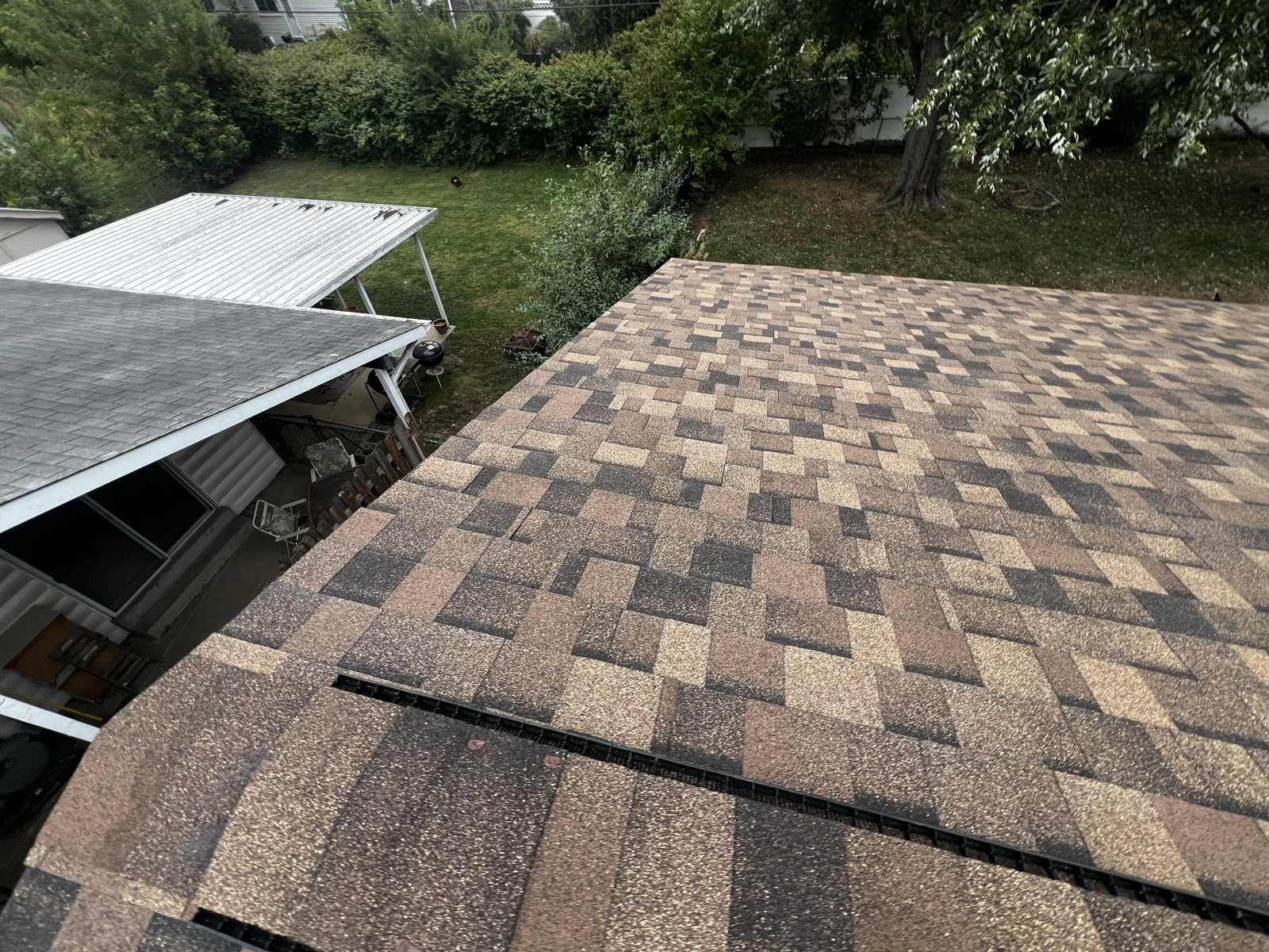 A wide plane of finished tan and brown architectural shingles meeting a ridge, with a carport and back garden beyond