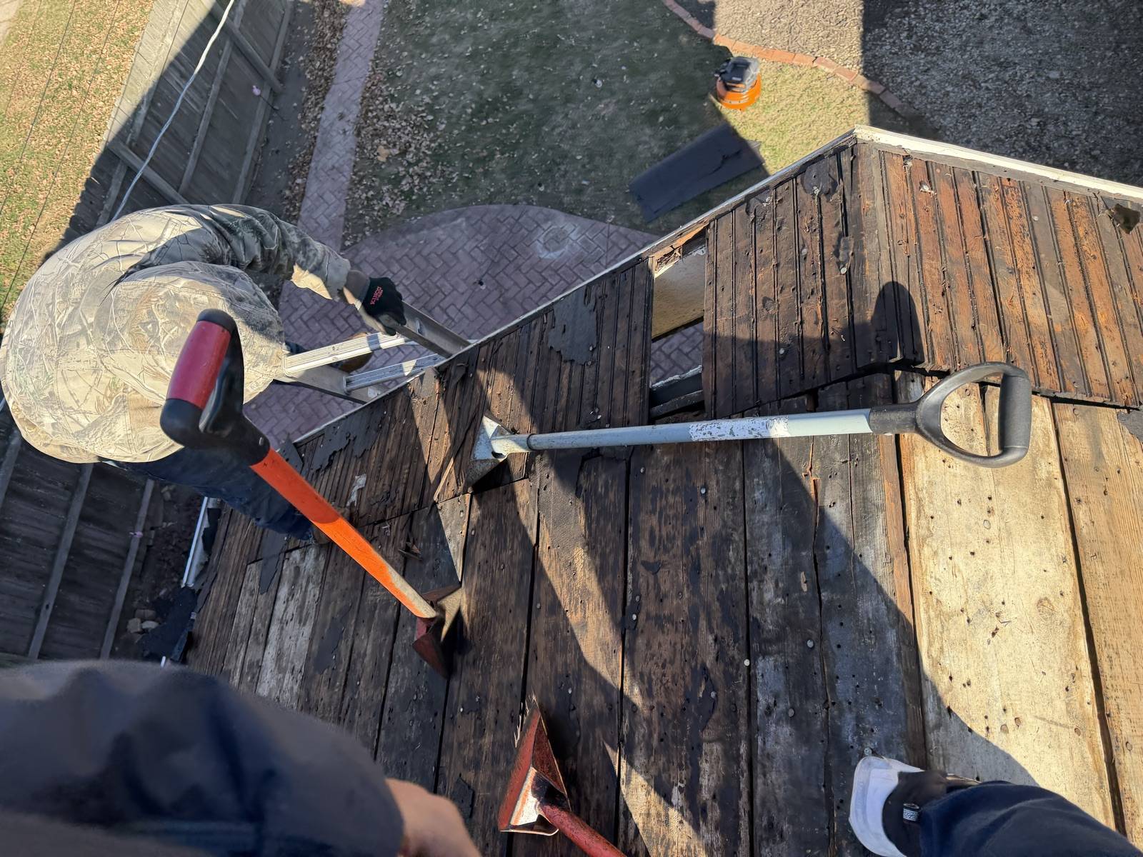 Two tear-off shovels lying on a stripped low-slope roof, one with a red handle and one with a steel shaft, with a roofer at the ladder and a brick patio below